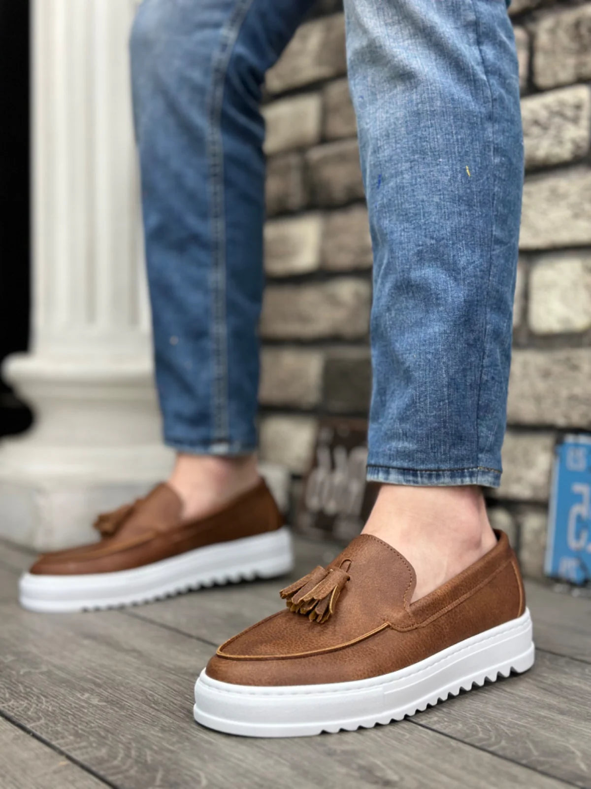 Brown loafers with tassels worn with blue jeans against a stone wall background